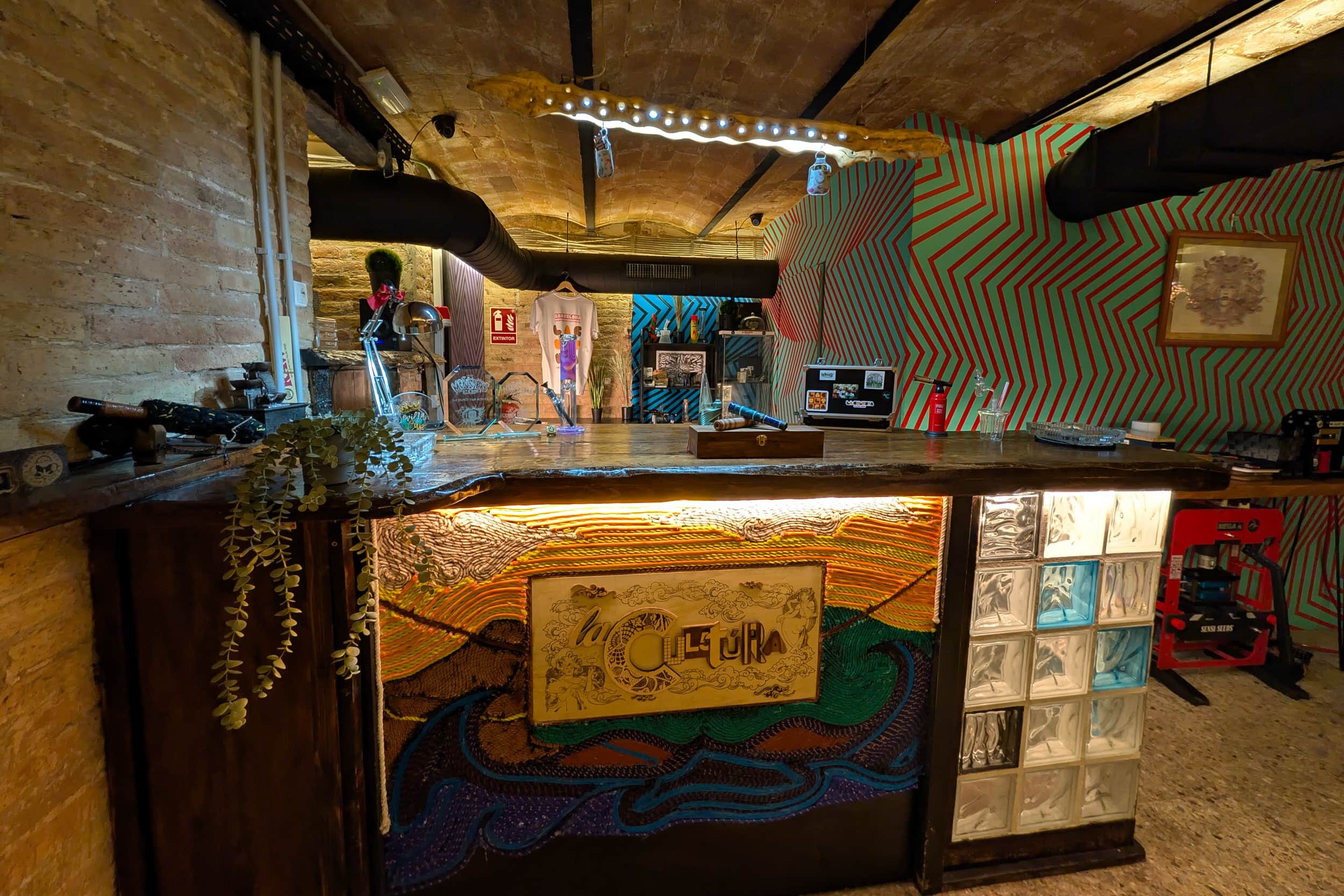 Zona de barra del club social cannábico La Cultura con mostrador artístico y pared vibrante en Barcelona